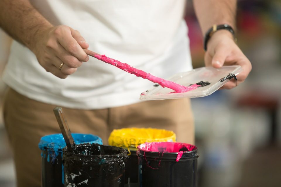 Händer blandar rosa tryckfärg med en spatel ovanför burkar med cyan, gul och magenta